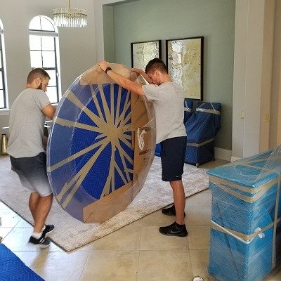 dinning area of a house. Movers packing a round table with plastic wrap, card board and tape.